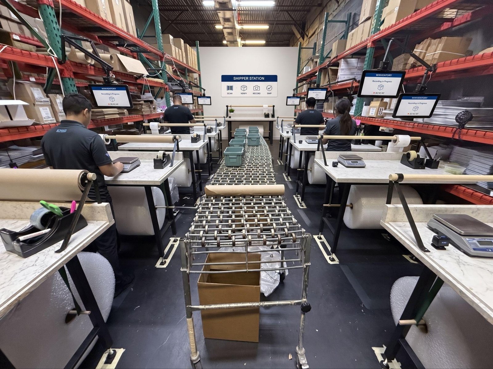 Two facing rows of packing benches with mounted screens, scales, wrapping material rolls and a roller conveyor down the centre aisle.