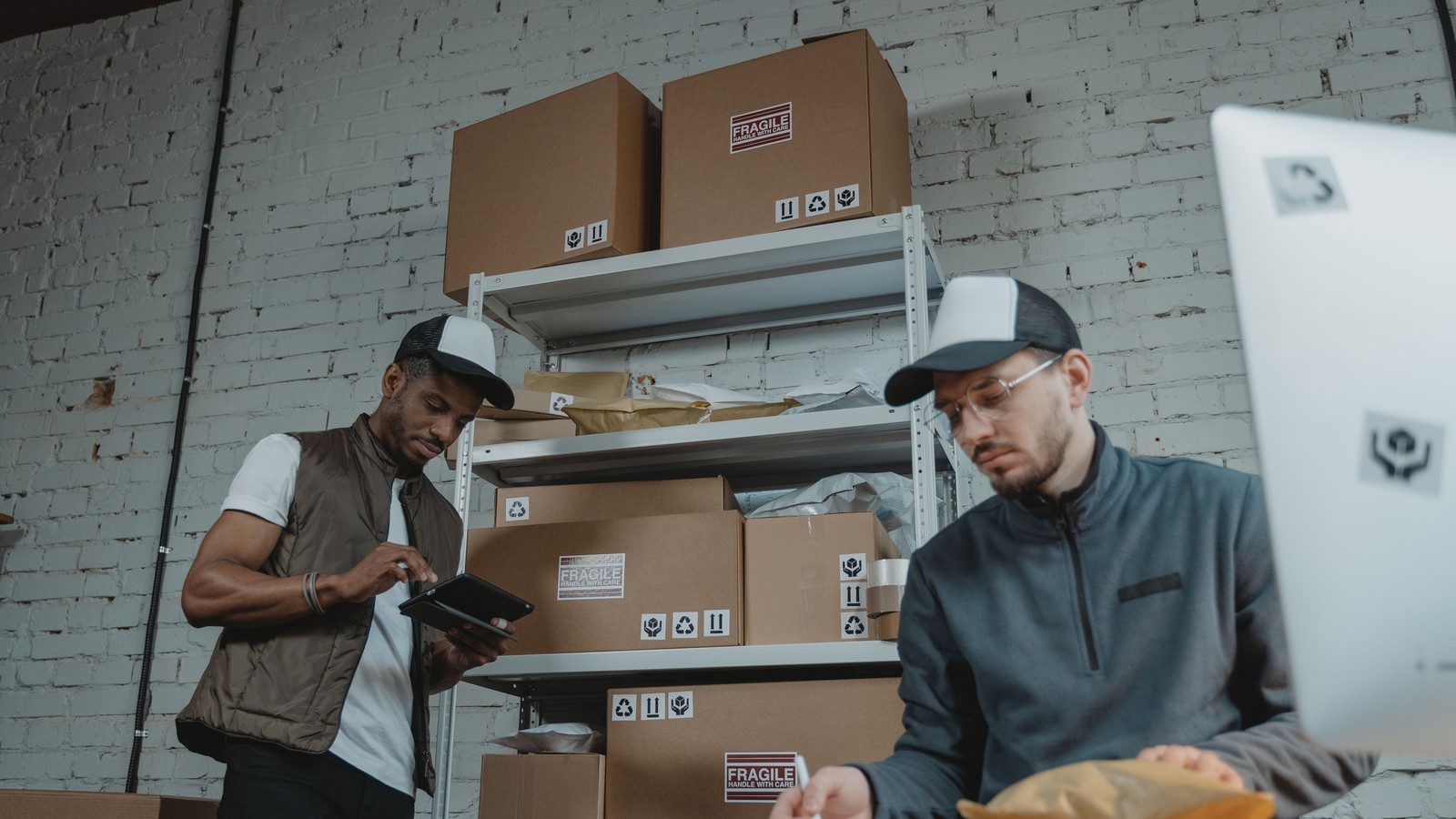 Two warehouse workers packing and checking orders on a tablet beside shelves of boxes