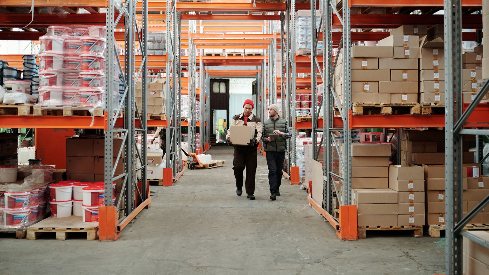 Two workers walking a warehouse aisle, one carrying a box and one holding a tablet