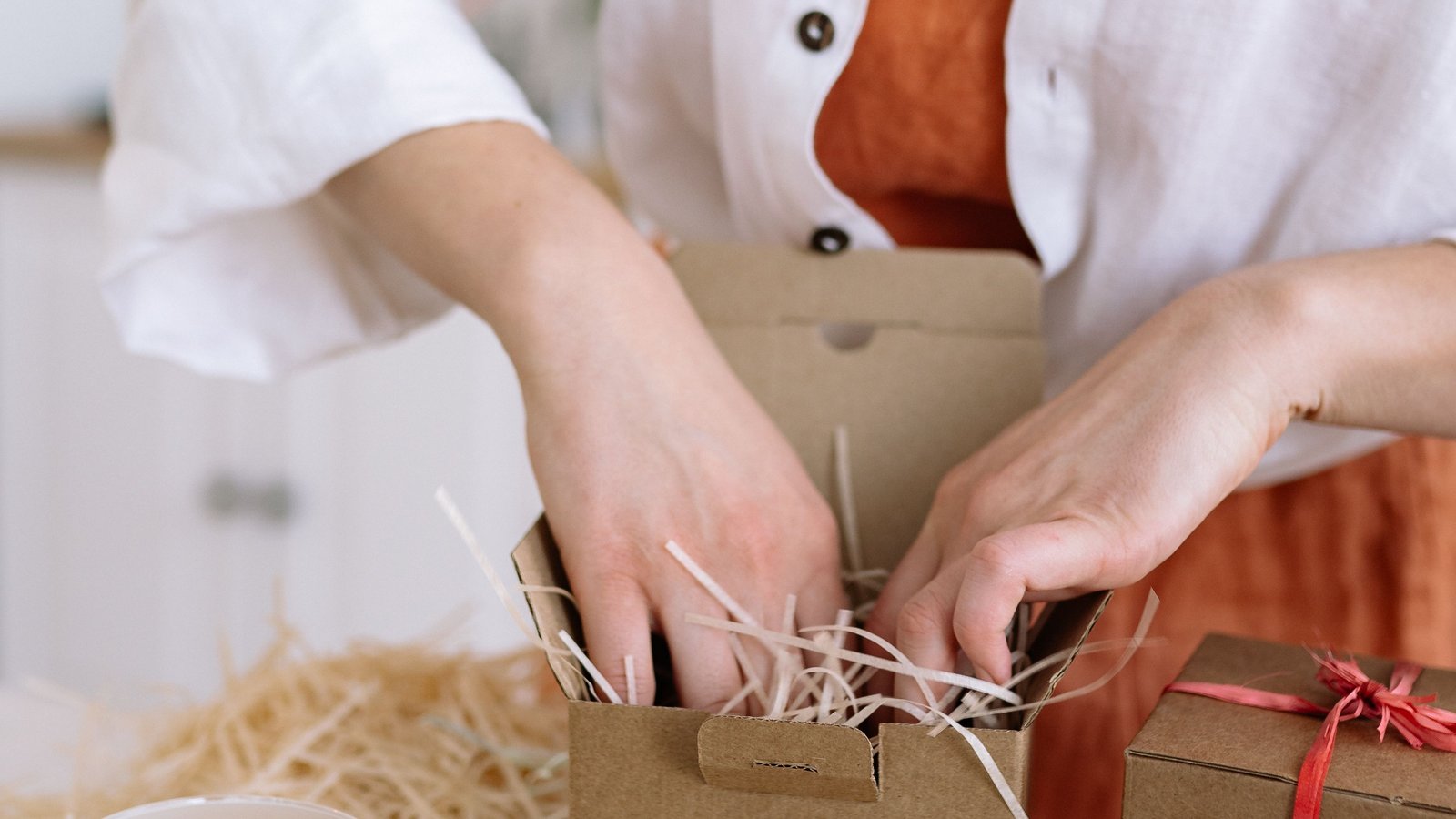 Hands filling a small kraft gift box with paper shred beside candles and a ribboned box