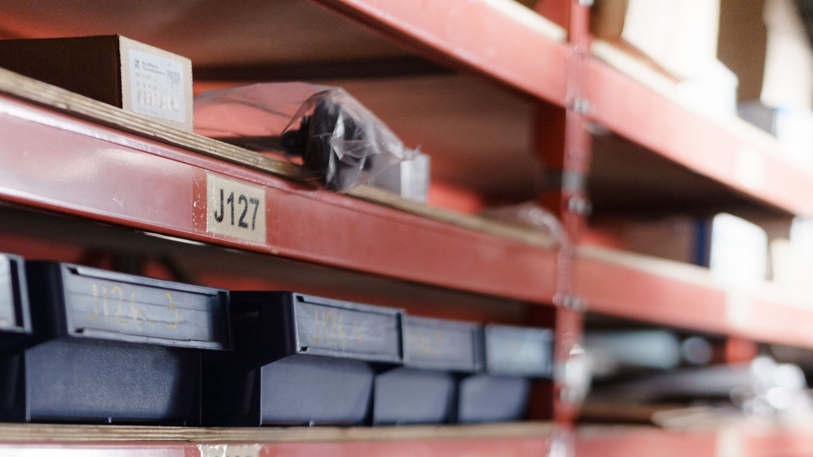 A hand pulling a small box from a labelled parts bin on red warehouse shelving