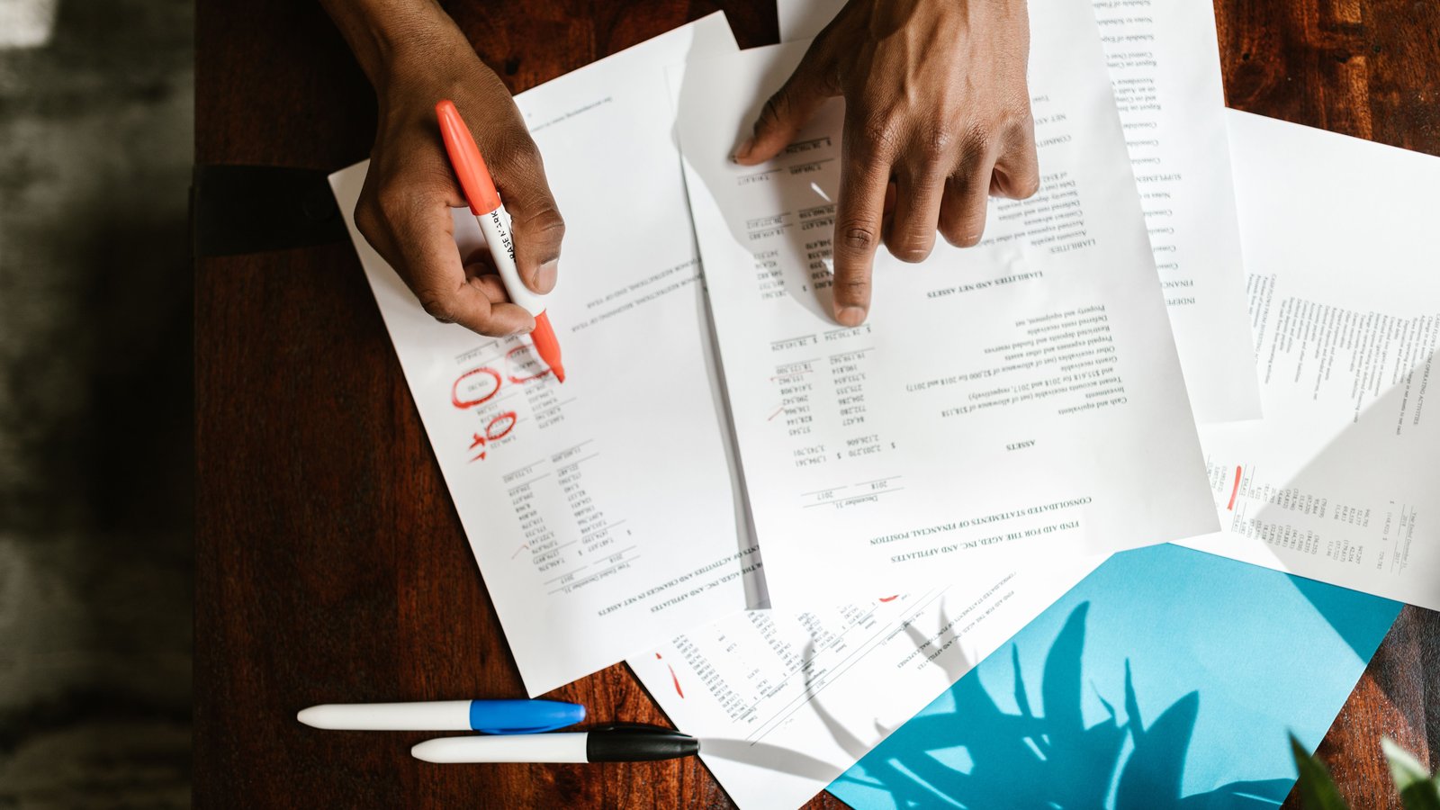 Hands marking up printed financial statements on a desk with coloured pens