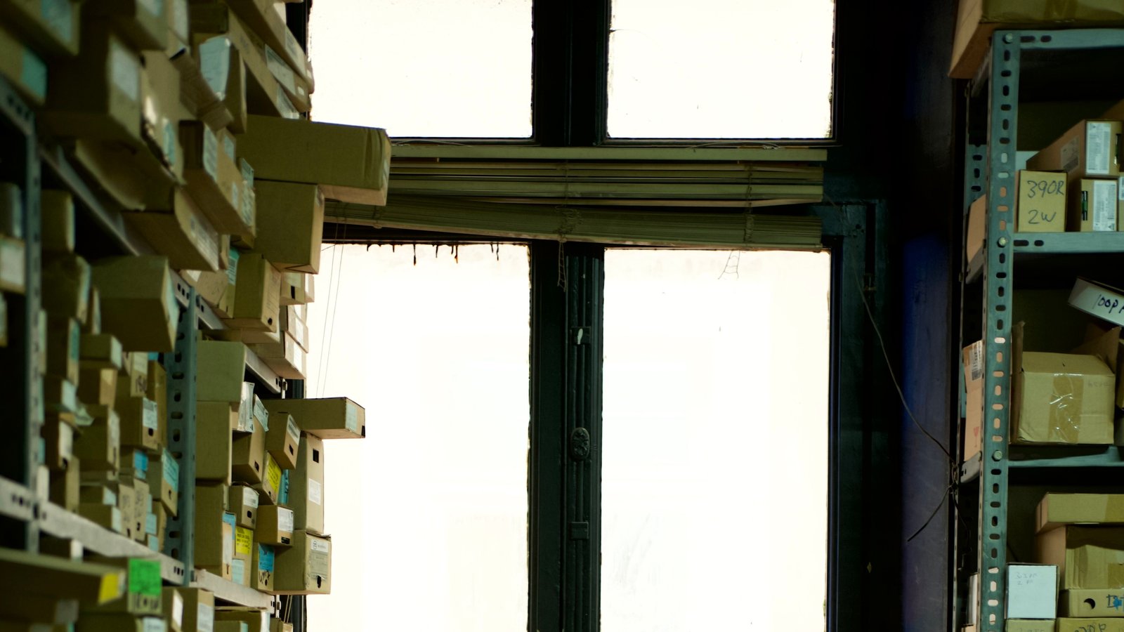 A crowded stockroom corner where boxes are stacked on shelving and piled on the floor by a window