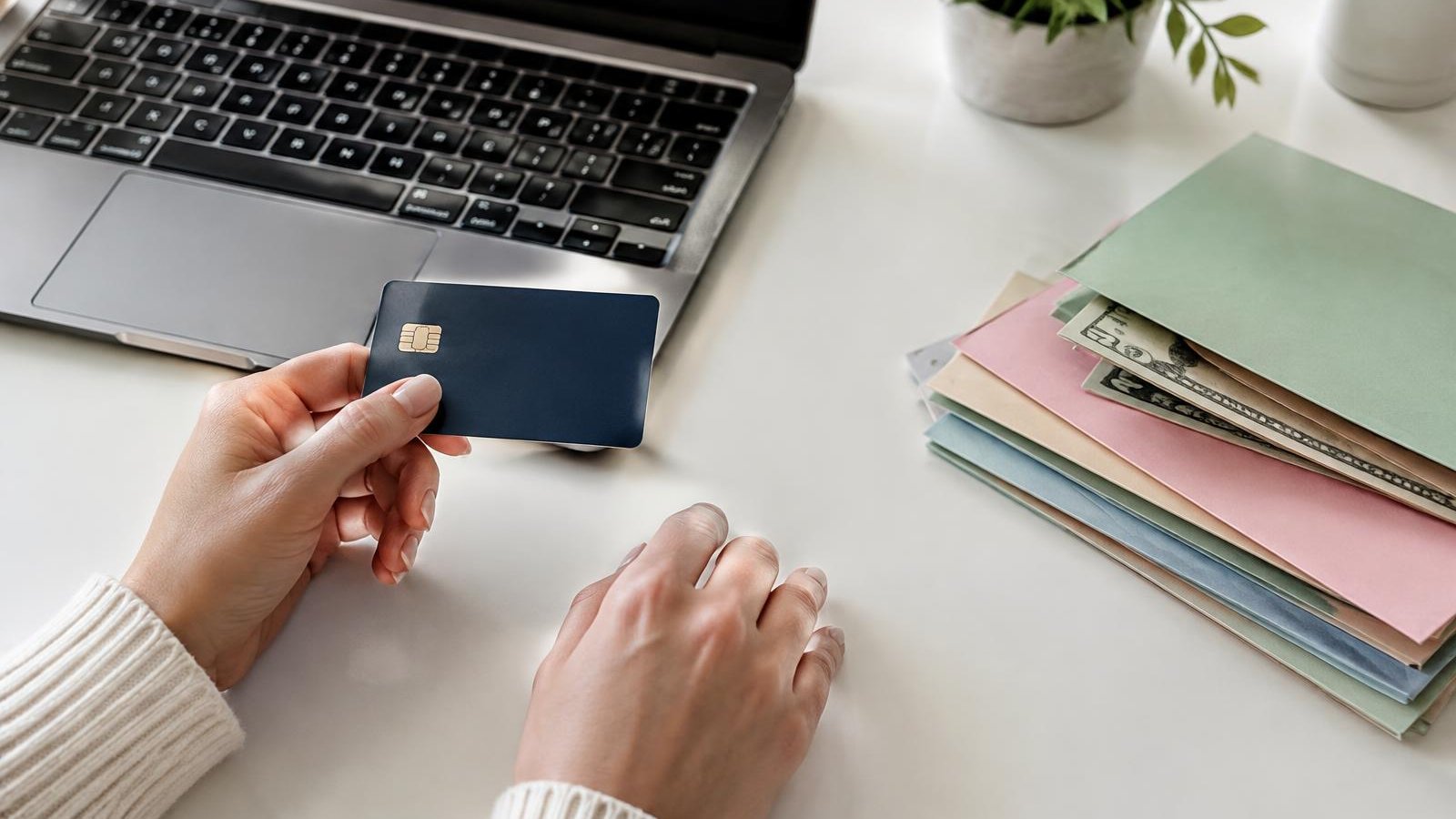 Hands holding a bank card beside a laptop and a stack of envelopes